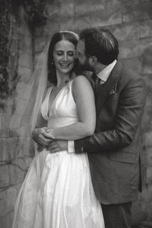 Black and white photo of a groom kissing his bride on the cheek as she laughs joyfully in her wedding dress.