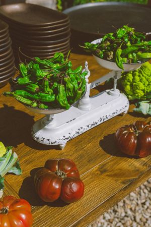 Vintage white balance scale holding fresh green peppers beside heirloom tomatoes and romanesco on a rustic wooden table.