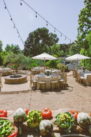 Outdoor wedding reception with round tables, white umbrellas, string lights, and fresh vegetables in a garden setting.