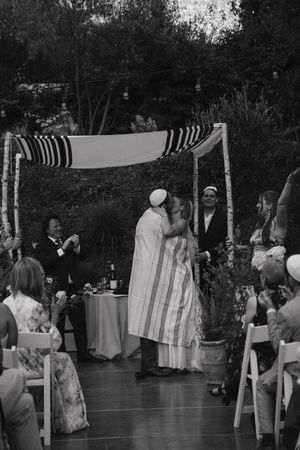 Black and white photo of a couple sharing a kiss under a chuppah at an outdoor Jewish wedding ceremony.