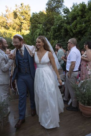 Joyful bride in white gown and groom in blue suit walk through cheering guests at outdoor wedding.