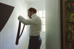 Bearded man in white shirt holding a brown tie while getting dressed near a bright window.