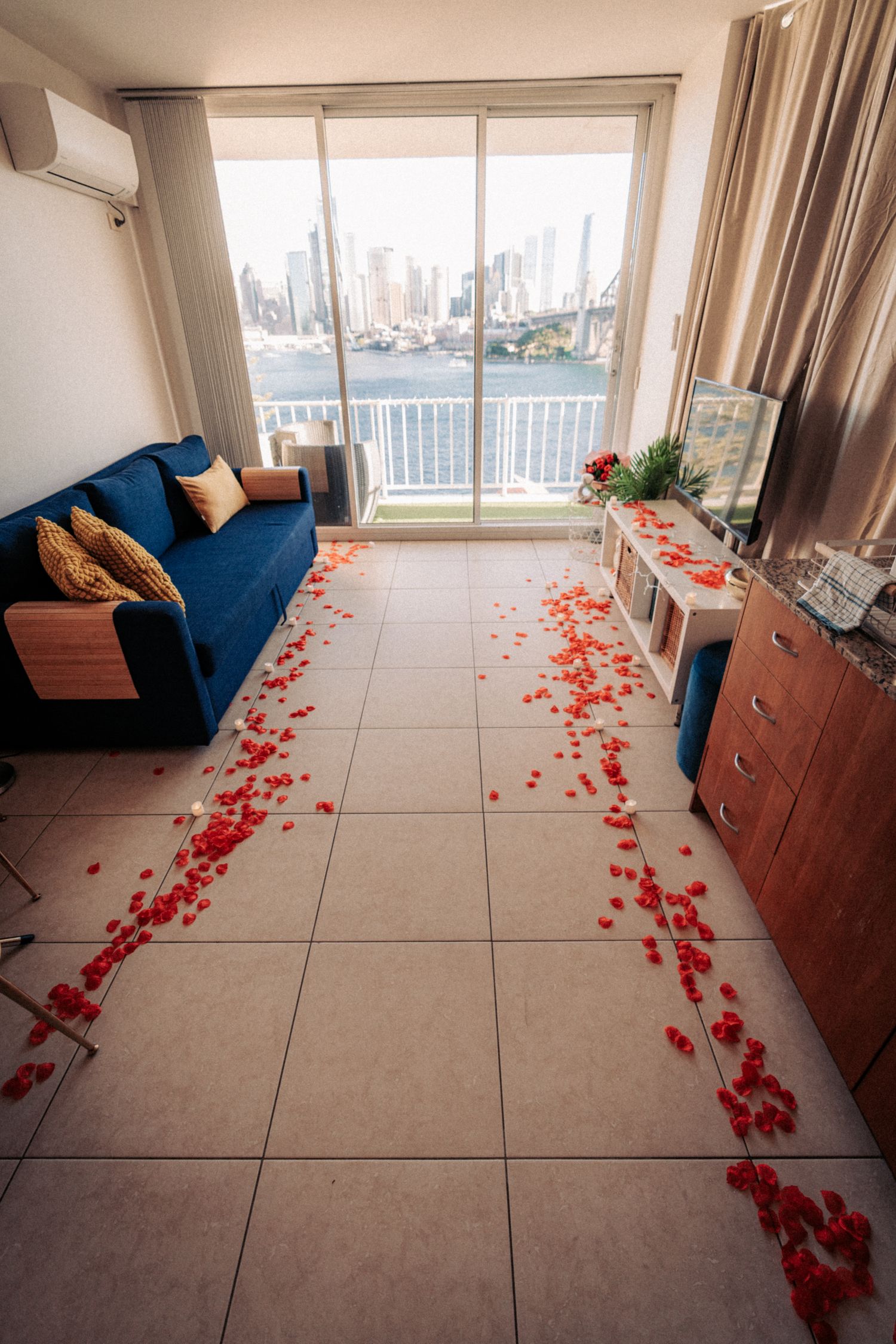 Romantic hotel room with red rose petals on tiled floor, blue sofa, and city skyline view through glass doors.