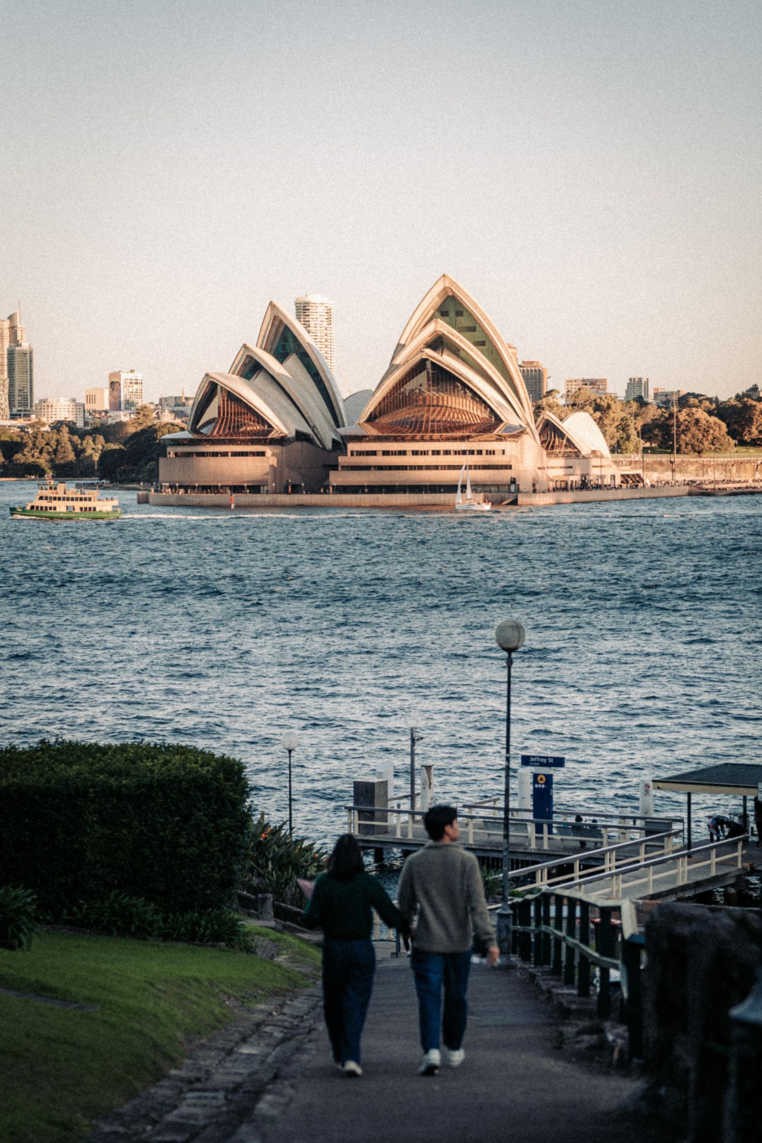 Couple holding hands walking toward Sydney Opera House waterfront view at golden hour in Australia.