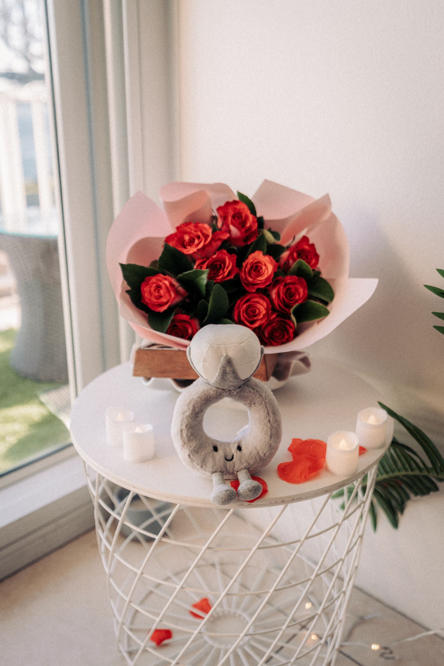Red rose bouquet in pink wrapping beside a stuffed toy, candles, and rose petals on a white wire table near a window.
