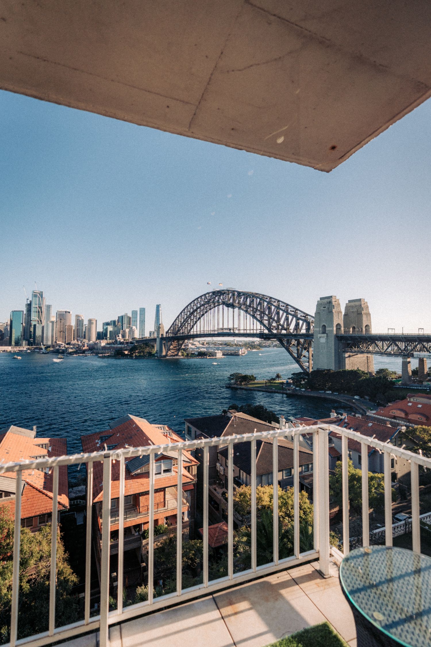 Balcony view of Sydney Harbour Bridge and city skyline on a clear sunny day, with blue waters and white railing.