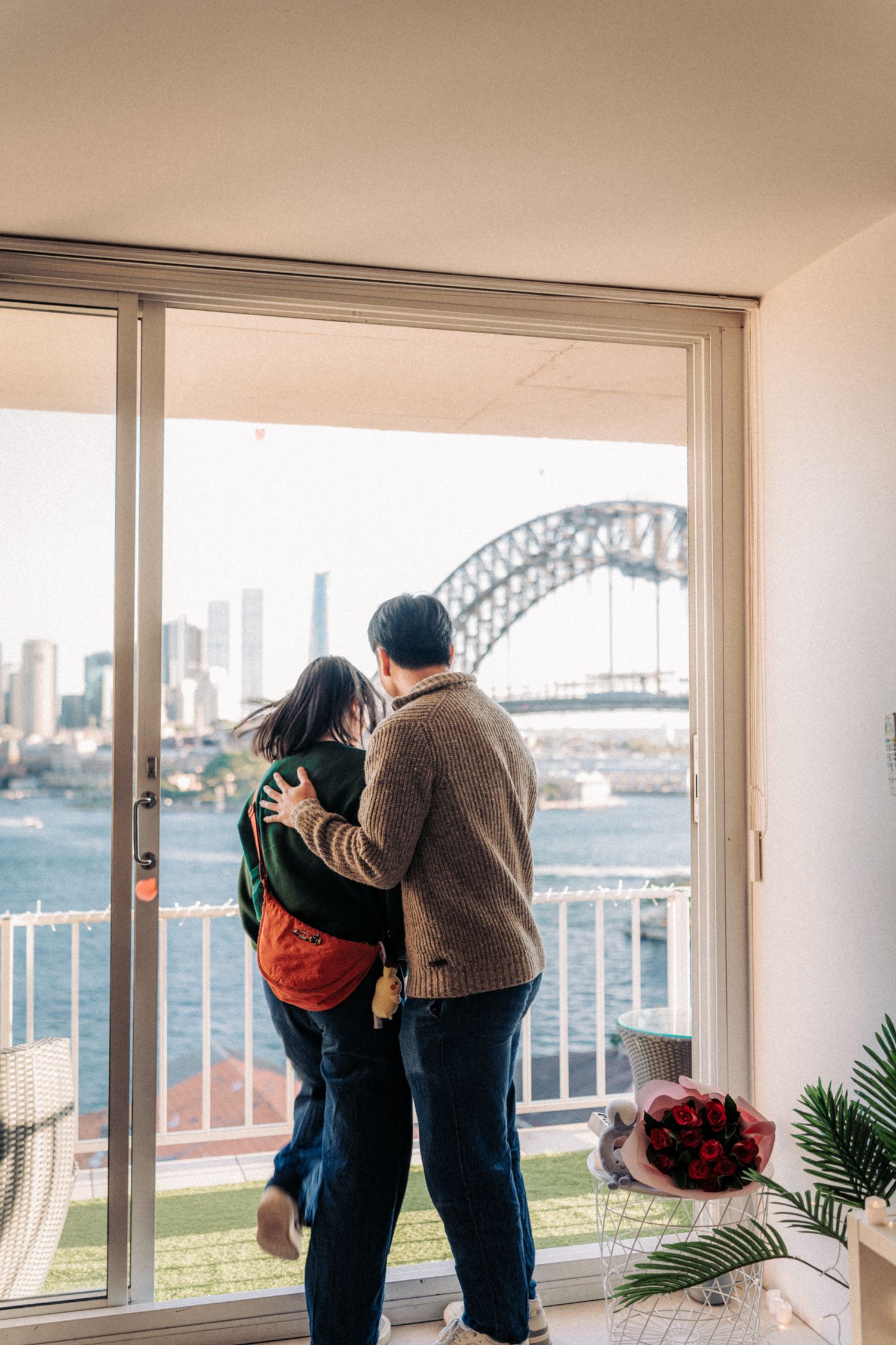 Couple embracing indoors with a stunning view of Sydney Harbour Bridge and city skyline through sliding glass doors.