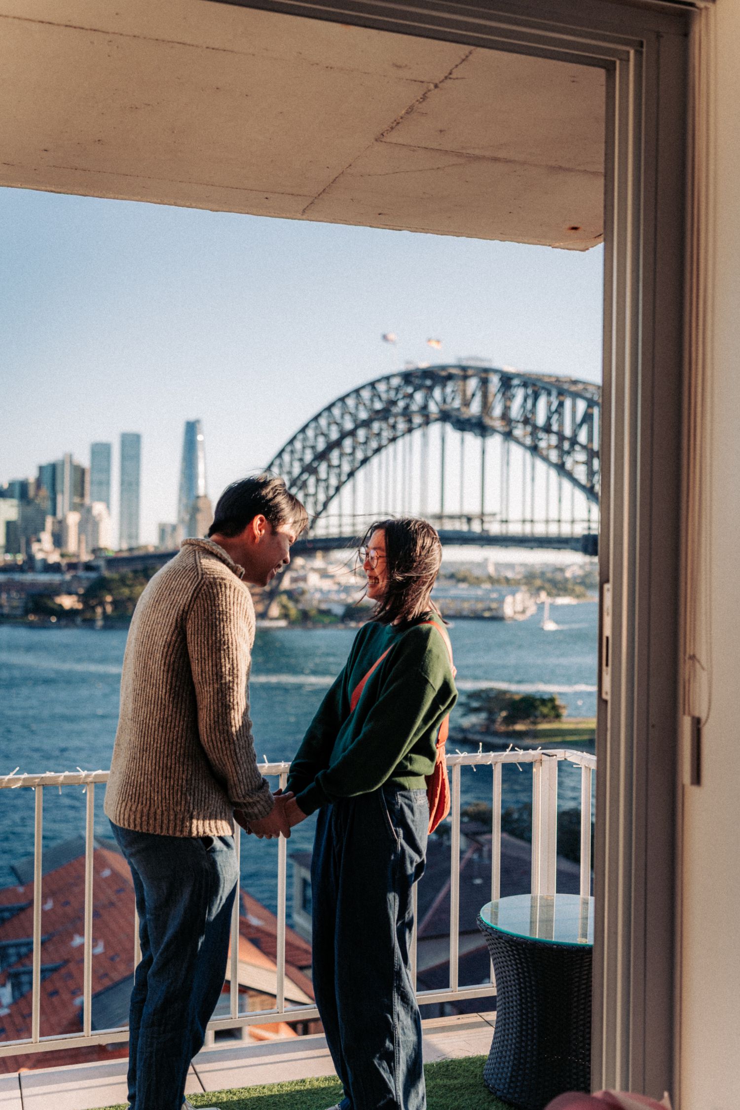 Couple holding hands on a balcony with Sydney Harbour Bridge and city skyline in the background.