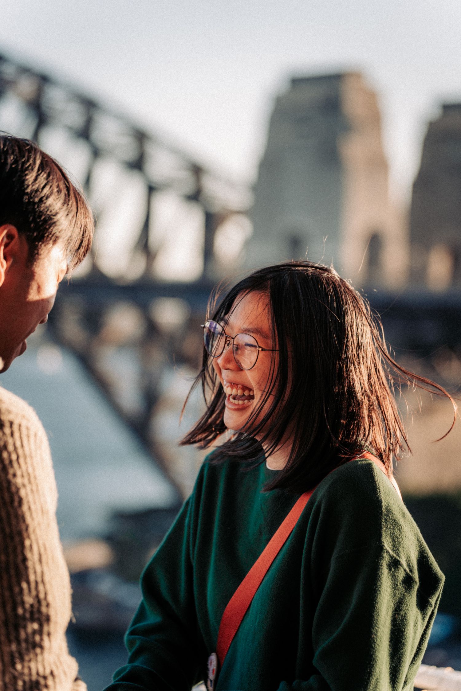 Young Asian woman in green sweater laughing near Sydney Harbour Bridge on a sunny day.