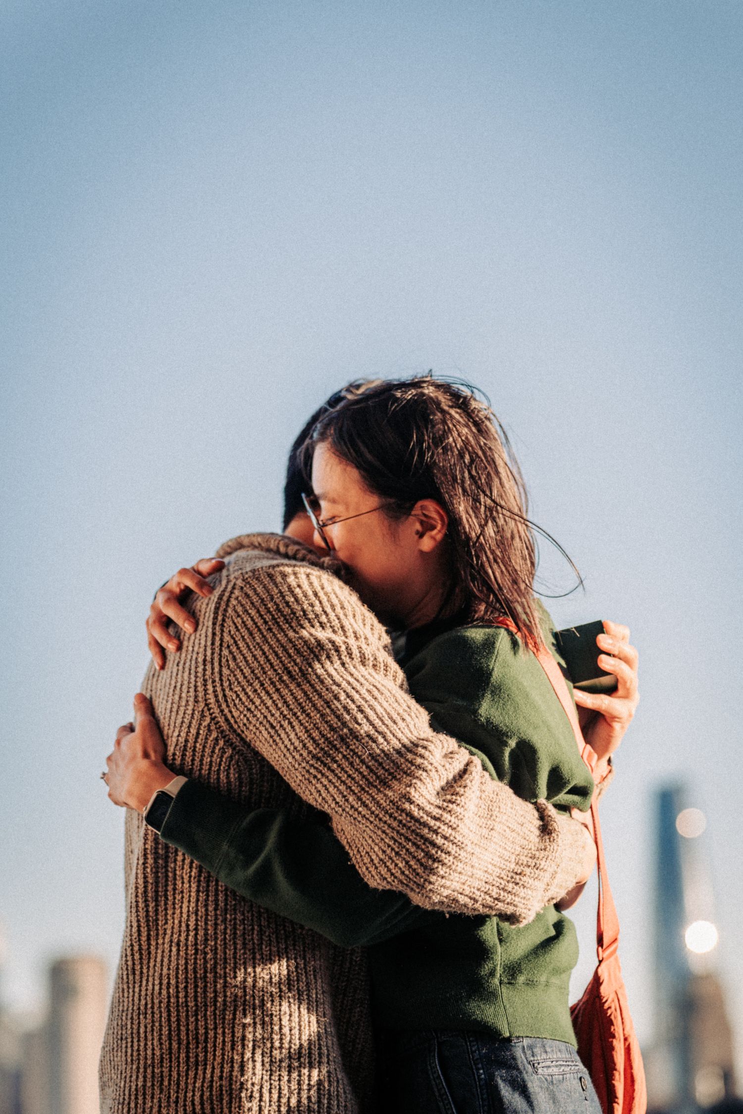 Two young women sharing a warm embrace outdoors with a city skyline in the background on a sunny day.
