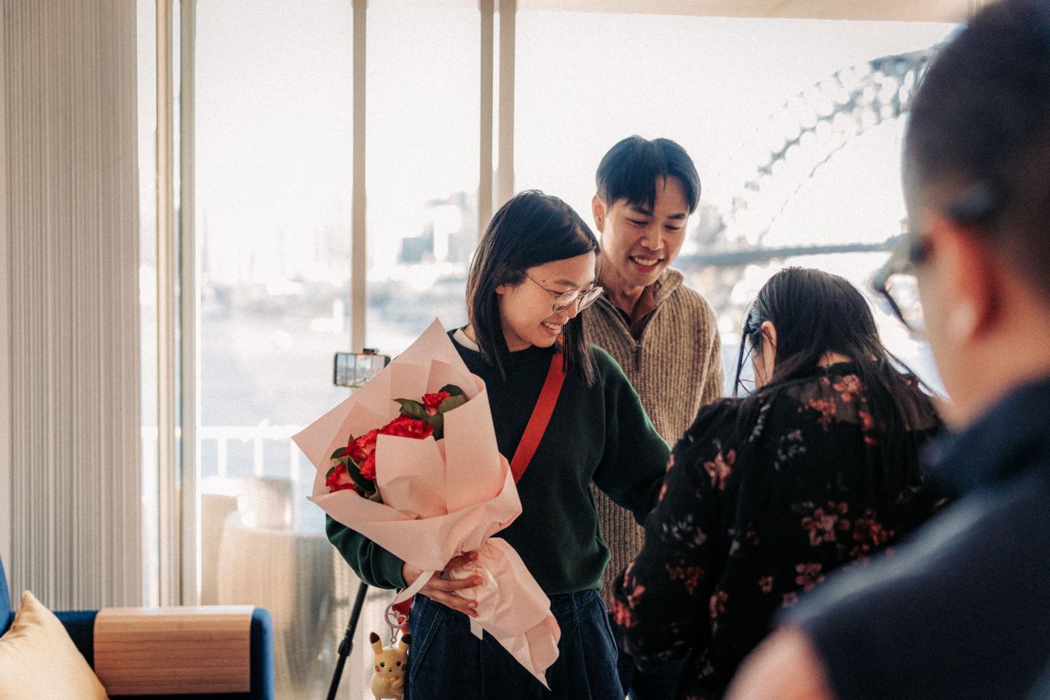 A smiling man presents a bouquet of red flowers to a woman at an indoor gathering near a large window.