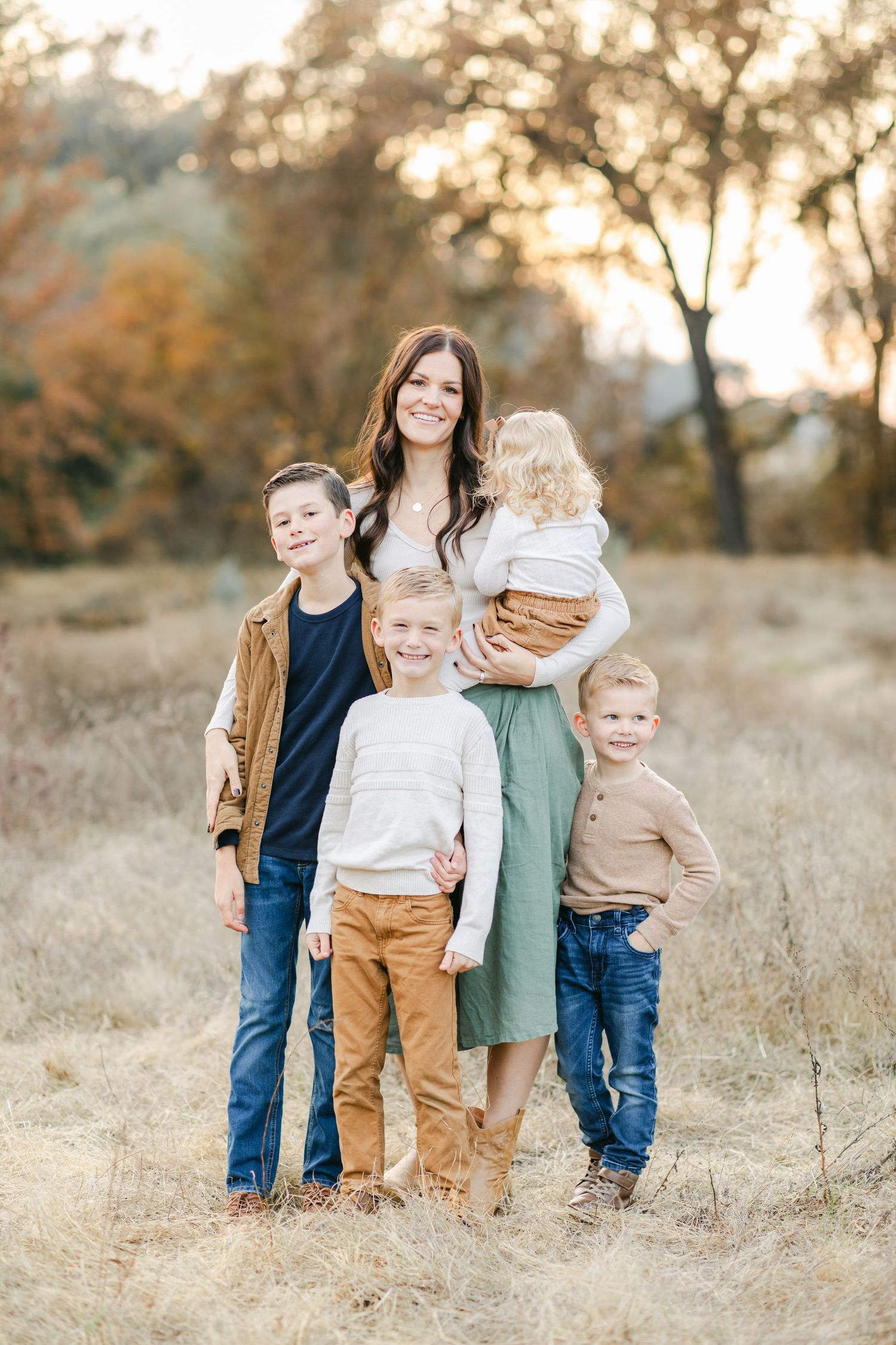 A smiling mother poses outdoors in a field with her four young children during a fall family portrait session.