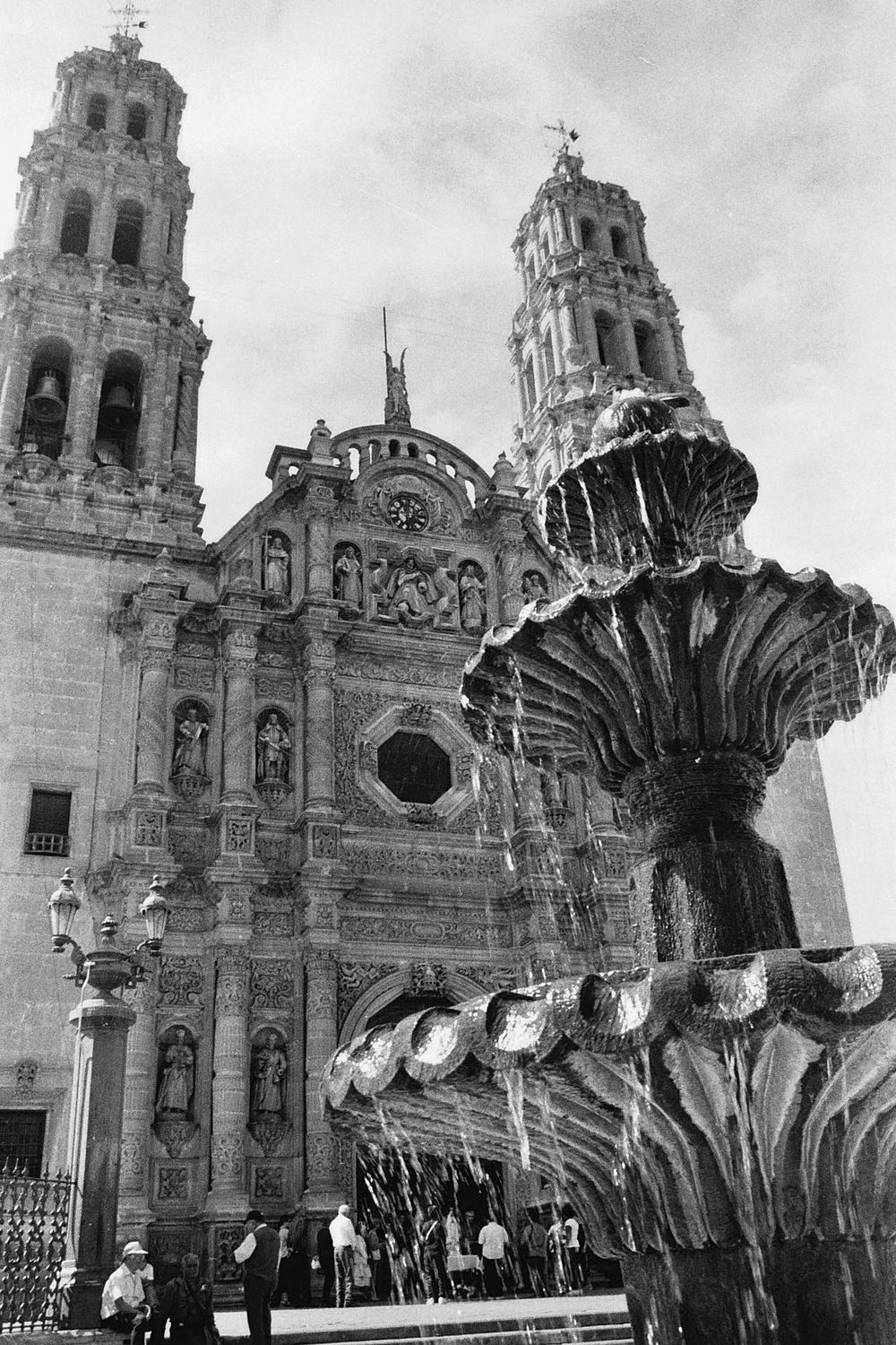Historic cathedral with ornate twin bell towers and decorative fountain in black and white.