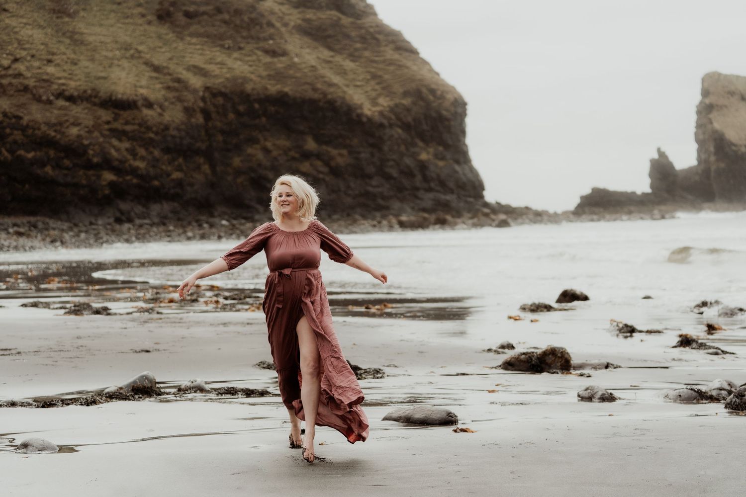 A woman in a flowing burgundy dress walks joyfully on a rocky beach with dramatic cliffs in the background.