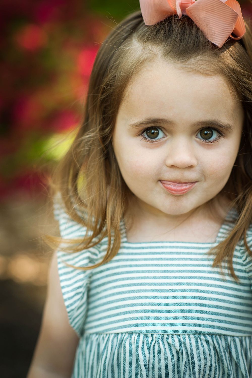 Adorable young girl with brown eyes and pink bow in hair wearing striped dress outdoors.