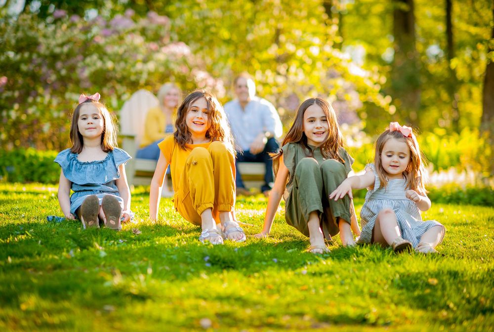 Four young girls sit on lush green grass in a sunny park with adults relaxing on a bench in the background.