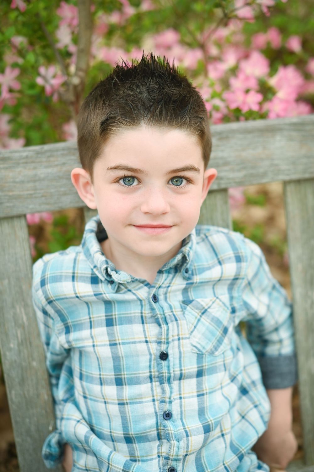Young boy with blue eyes wearing a plaid shirt, smiling on a wooden bench with pink flowers in background.