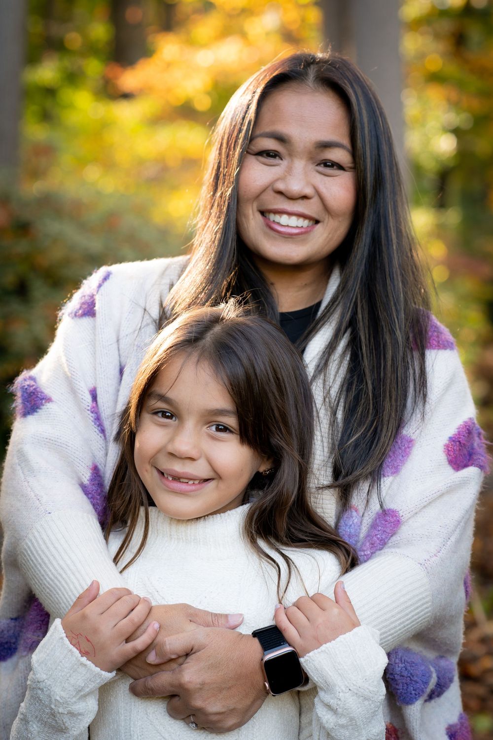 A smiling woman hugs a young girl outdoors in an autumn forest, both wearing cozy white sweaters.