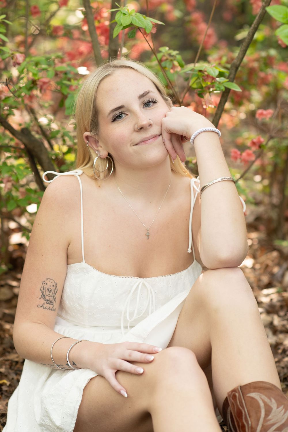 Young blonde woman in white sundress with tattoo and bracelets sitting outdoors among colorful autumn foliage.