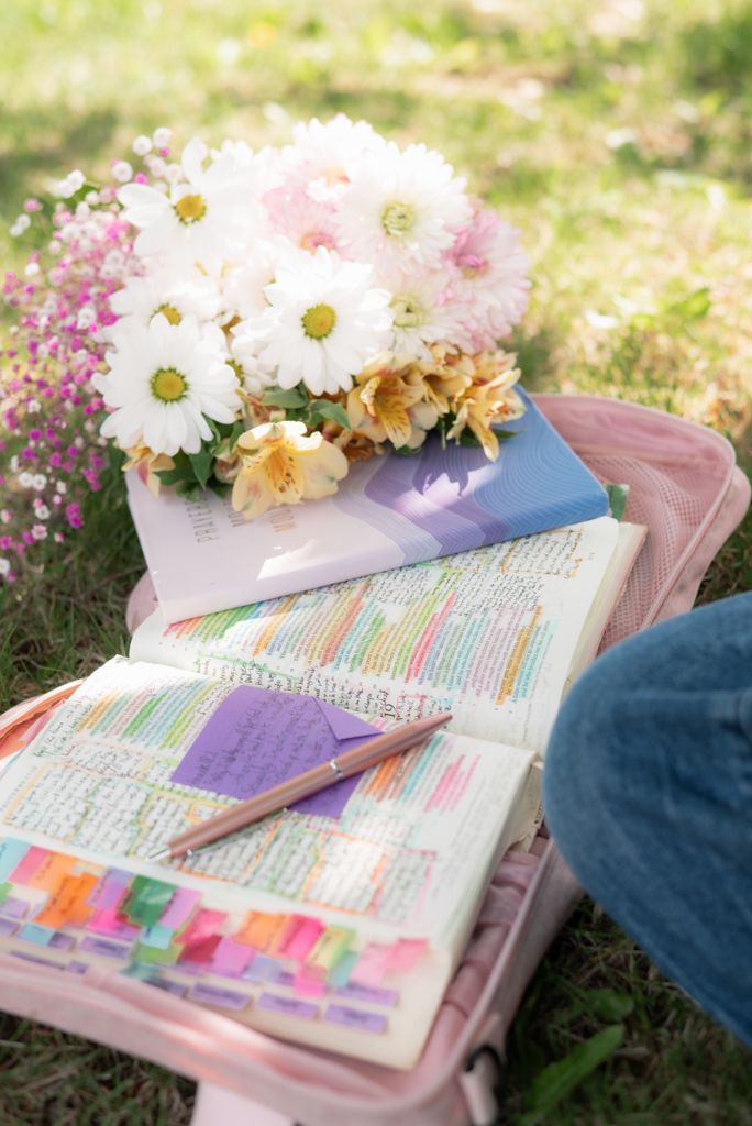 Open Bible with colorful tabs and sticky notes beside a bouquet of daisies on a pink bag outdoors.