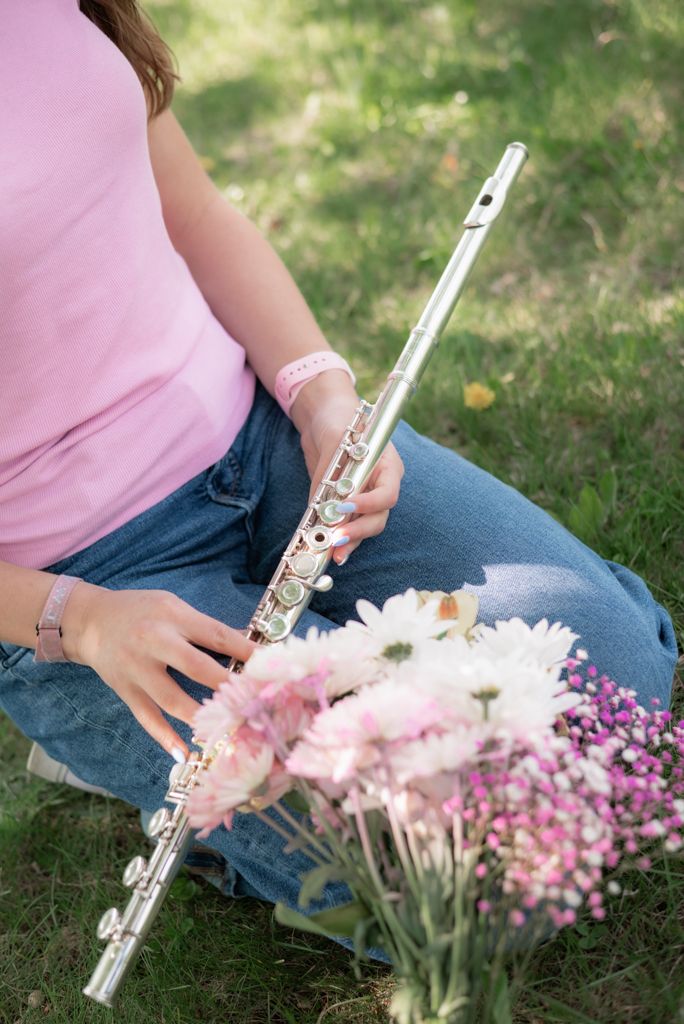 Girl in pink shirt holding a silver flute outdoors, sitting on grass with pink and white flowers nearby.