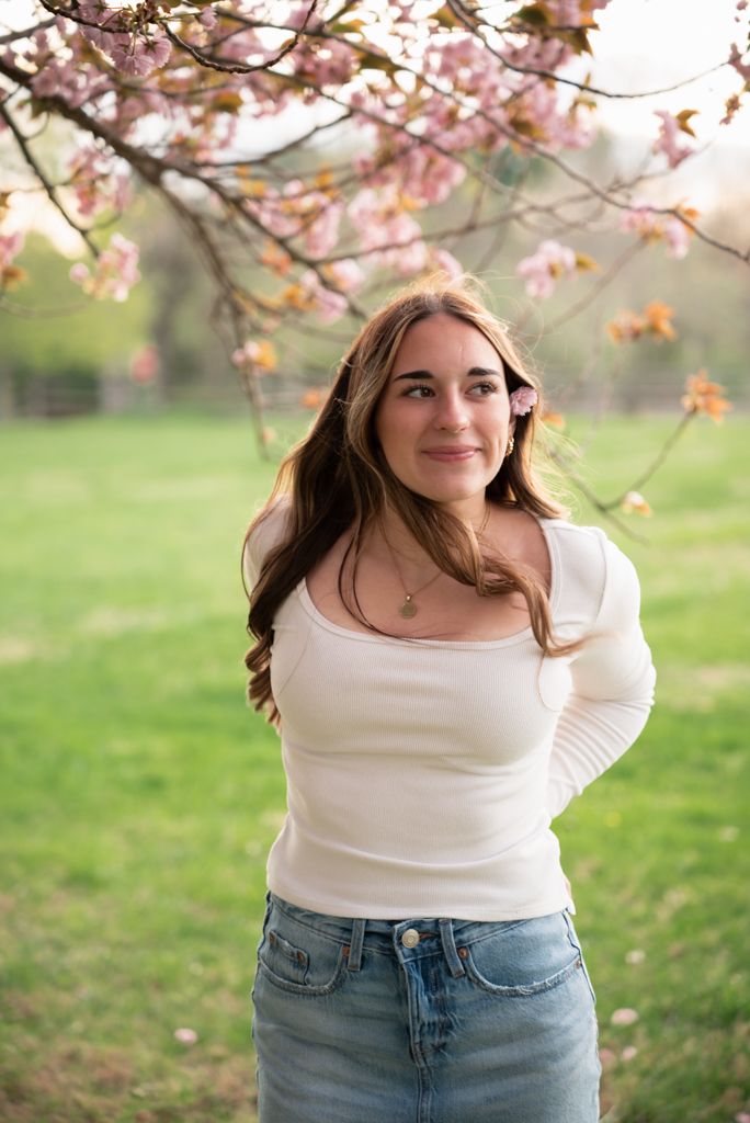 Young woman in white top and denim skirt poses under pink cherry blossoms in a lush green park.
