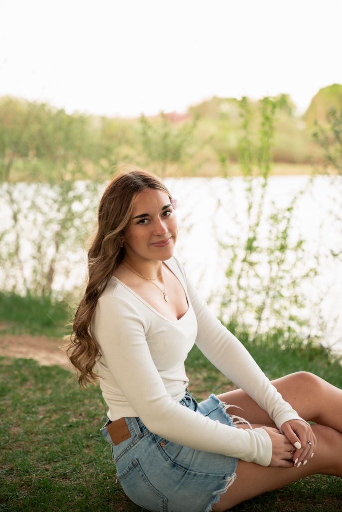 Young woman in white top and denim skirt sitting by a serene lake surrounded by lush green trees.