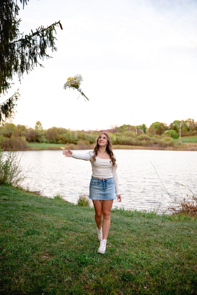 Young woman in denim skirt tossing a wildflower bouquet by a serene lake at golden hour.