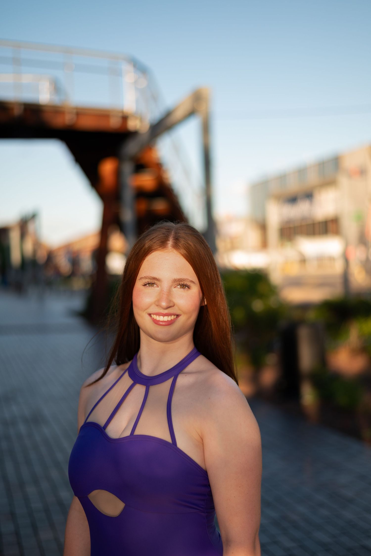 Smiling young woman in purple halter dress poses outdoors near industrial bridge structure in warm golden hour light.