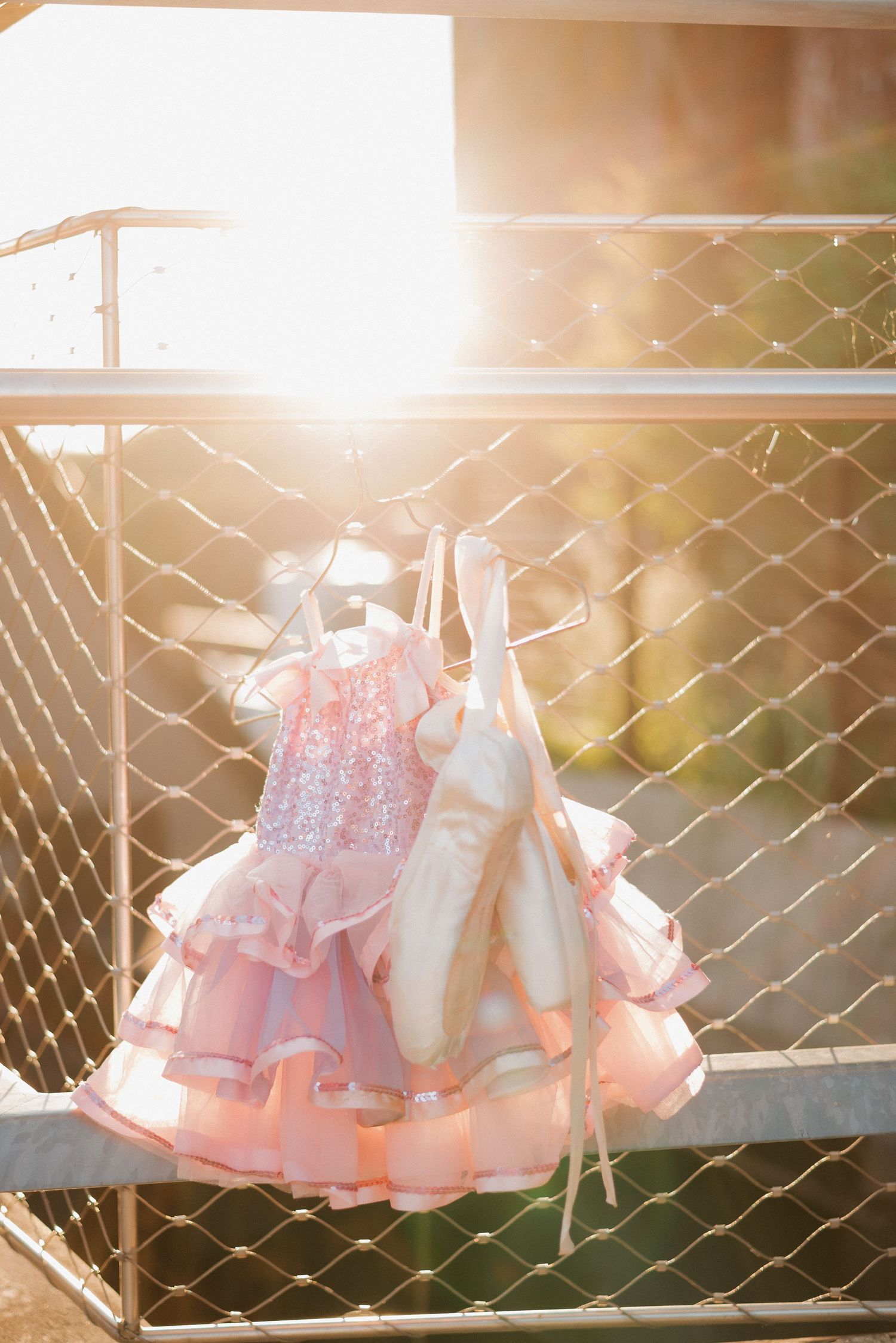Pink ruffled girls dress and ballet pointe shoes hanging on metal fence railing in golden sunlight.