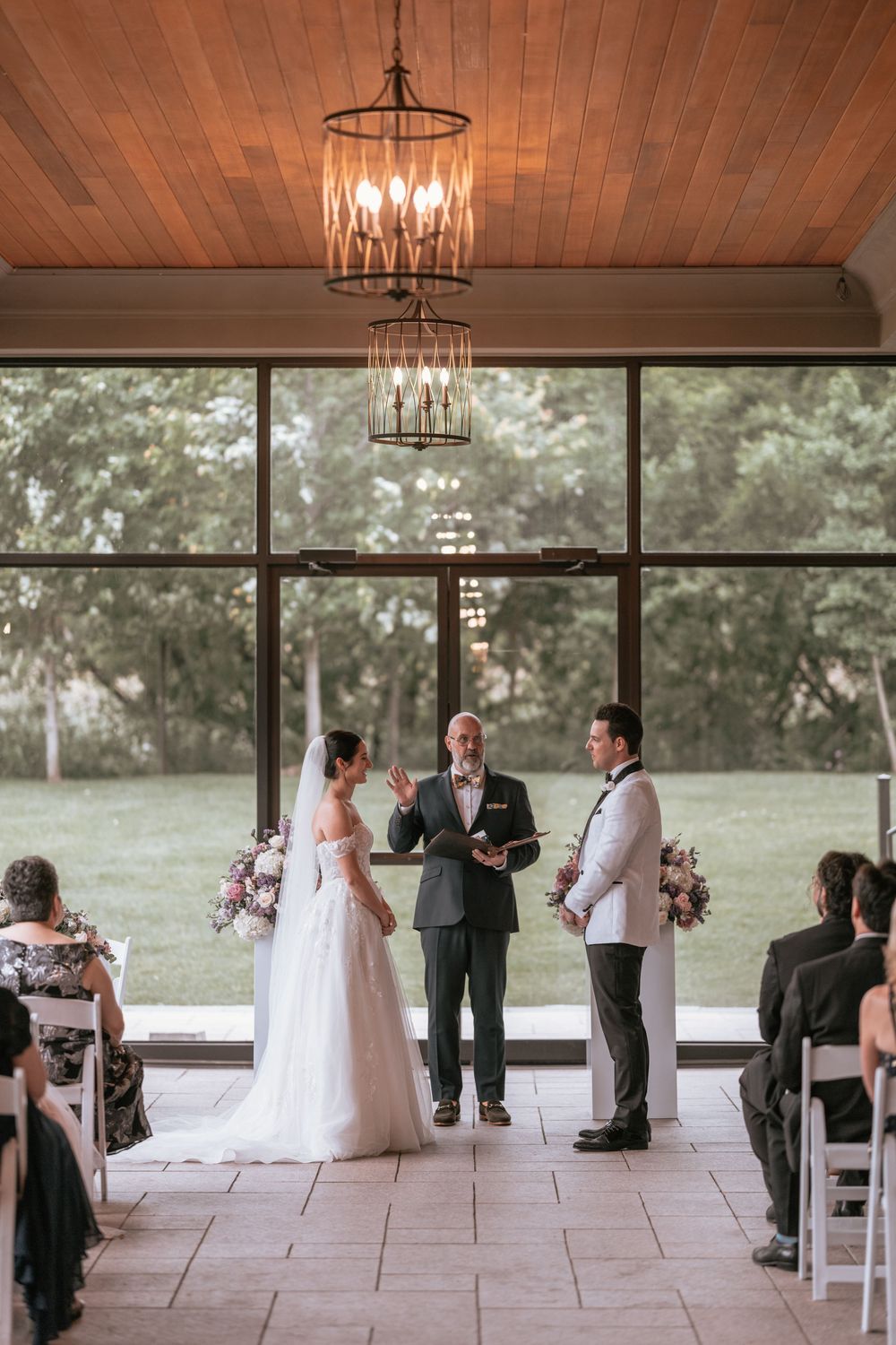 Bride and groom exchange vows at elegant indoor wedding ceremony with chandelier and floor-to-ceiling windows.