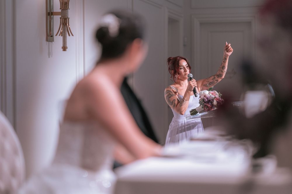 A tattooed bridesmaid gives a heartfelt speech gesturing expressively while the bride listens at a wedding reception.