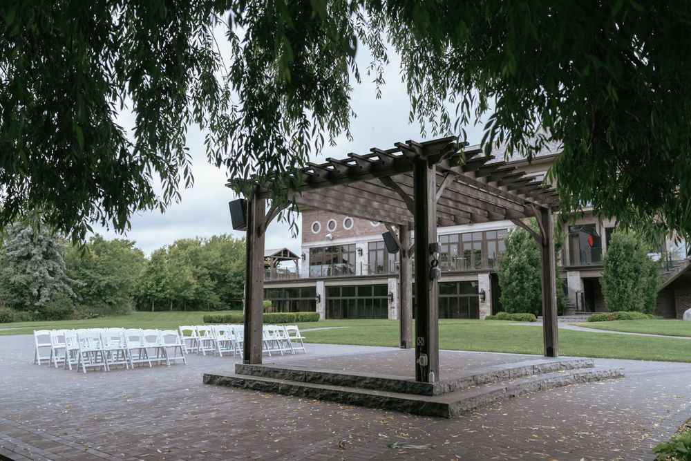 Outdoor wedding ceremony setup with wooden pergola, white chairs, and lush greenery near an elegant venue.