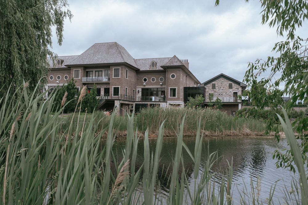 Brick mansion with balconies sits beside a calm pond, framed by tall reeds and weeping willow trees under cloudy skies.