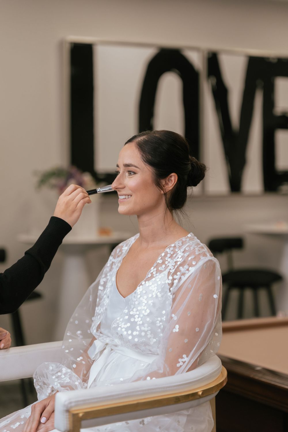Happy bride in white sheer robe smiling as makeup artist applies foundation with a brush in a bright salon.