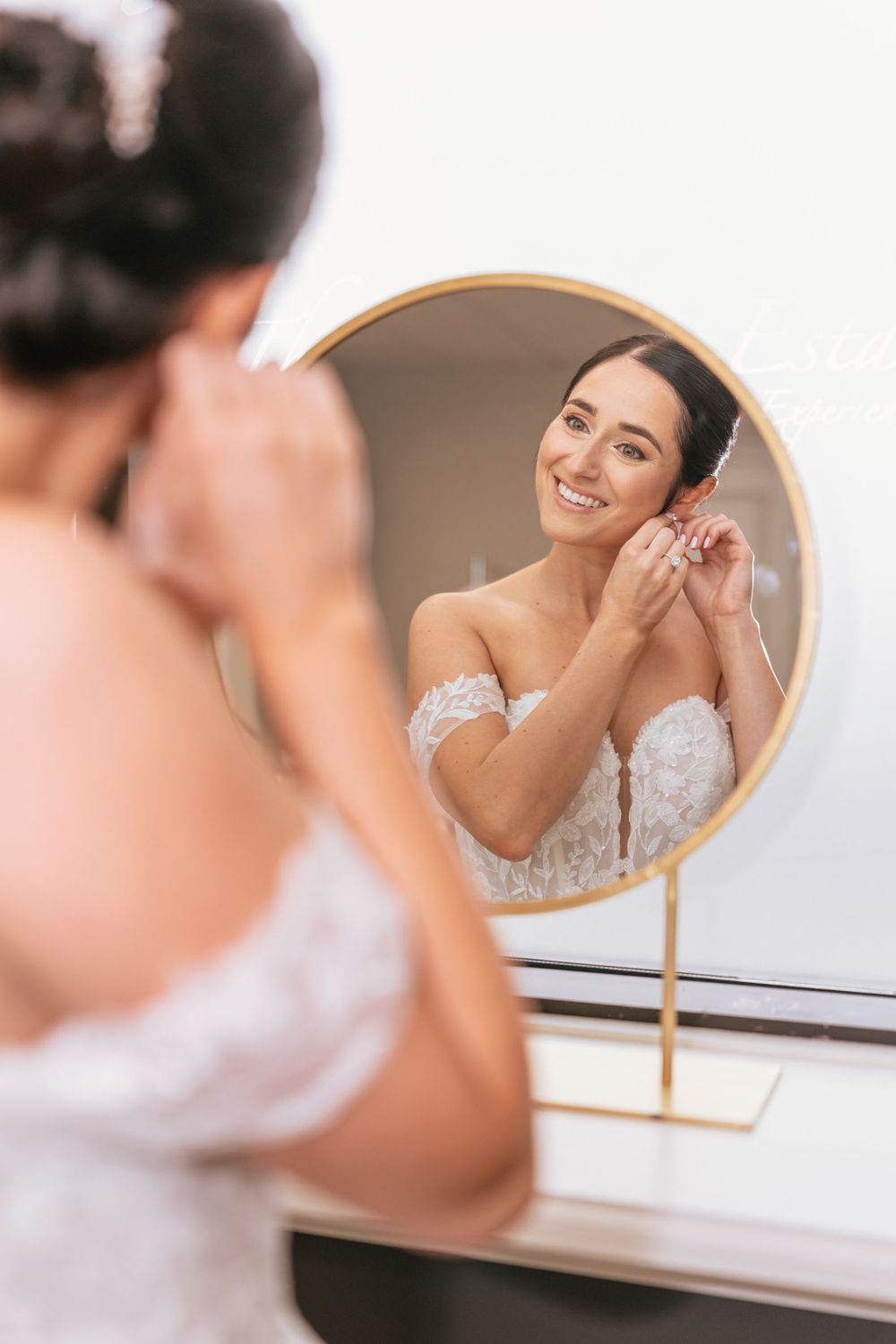 Smiling bride in lace off-shoulder gown putting on earrings while looking at her reflection in a round gold mirror.