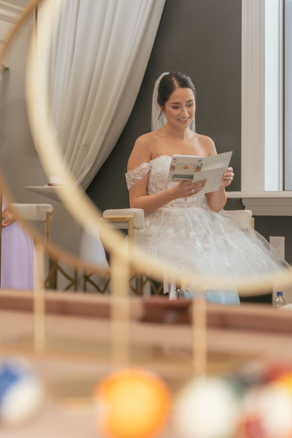 Bride in off-shoulder lace gown smiling while reading a card at her wedding ceremony.