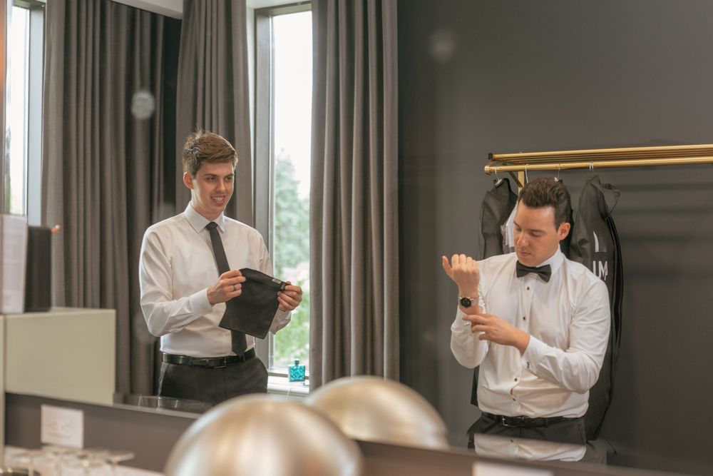 Two men in formal attire prepare for an event, one adjusting cufflinks and another holding a jacket.