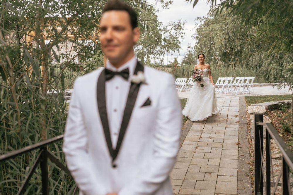 Groom in white tuxedo stands in foreground as bride in wedding gown approaches on outdoor pathway.