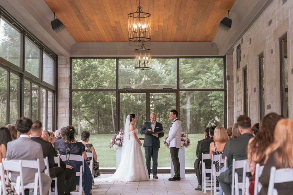 Elegant indoor wedding ceremony in a sunlit venue with large windows, white chairs, and a couple exchanging vows before guests.