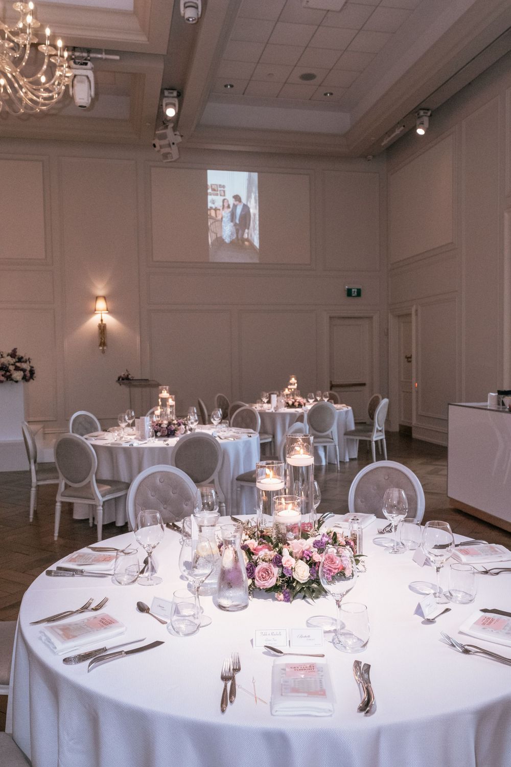 Elegant wedding reception hall with round tables, white linens, pink floral centerpieces, candles, and a chandelier.