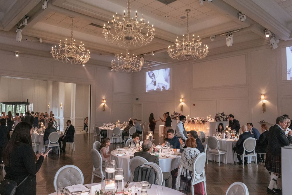 Elegant wedding reception hall with crystal chandeliers, round white tables, and guests dining beneath a projected photo display.