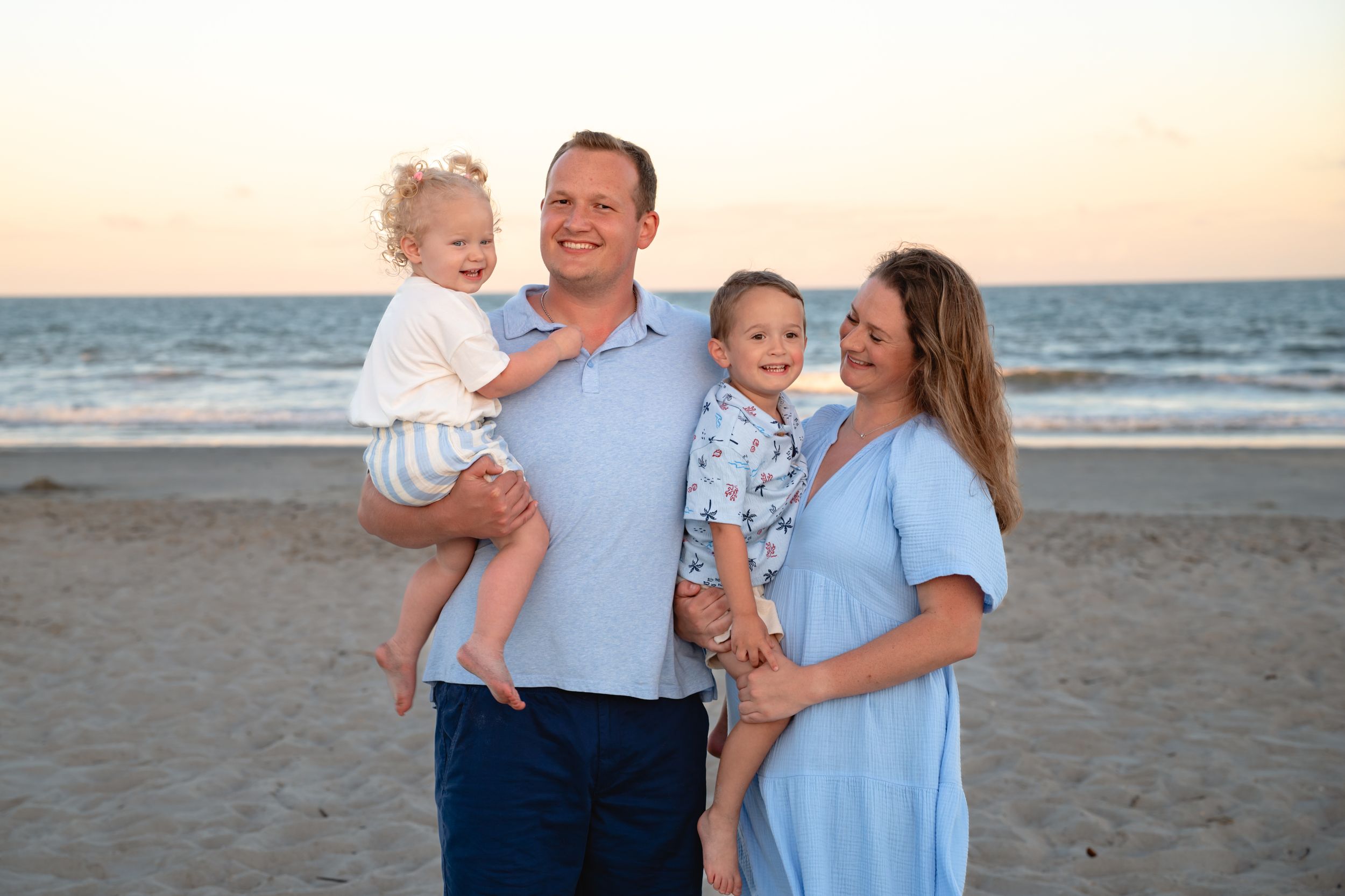 Family of four smiling on a sandy beach at sunset, parents each holding a young child in light blue outfits.