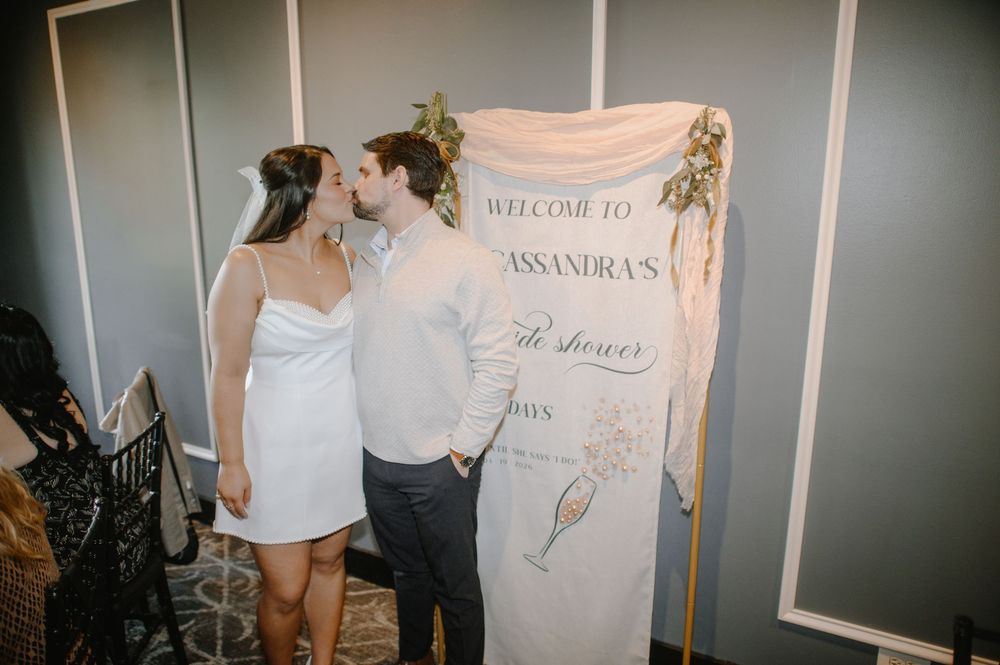 Couple kissing at a bridal shower welcome banner, bride in white mini dress, groom in gray suit.