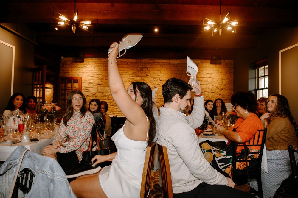 Bride and groom joyfully wave white fans at their wedding reception in a rustic brick-walled venue with guests dining.