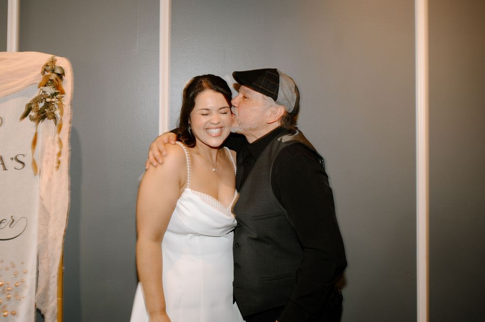 Bride in white dress receives a kiss on the cheek from an older woman in black at a wedding celebration.