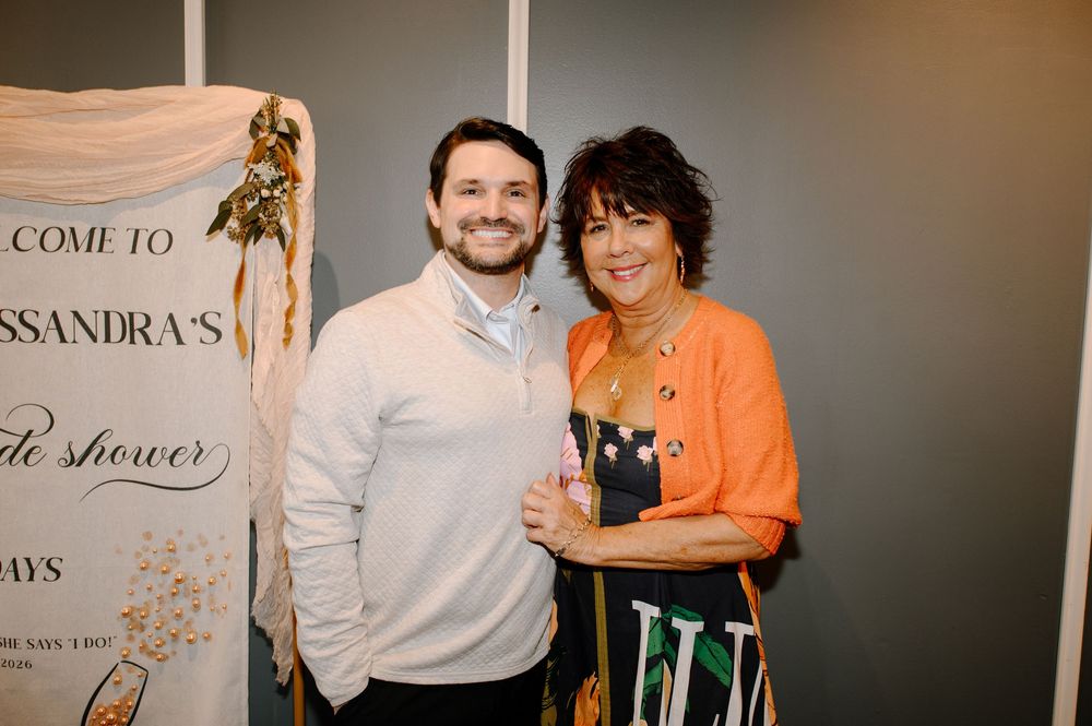 A man and woman smiling together at a bridal shower event, posing in front of a decorative banner.