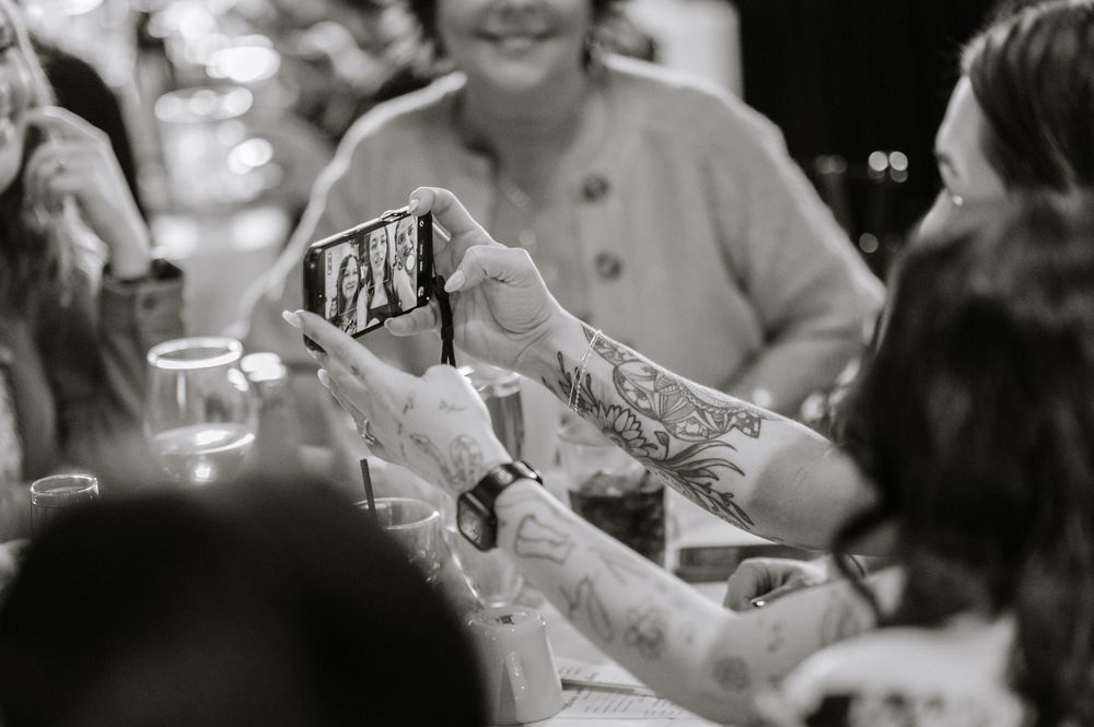 Black and white photo of tattooed arms holding a smartphone at a social gathering or wedding reception.
