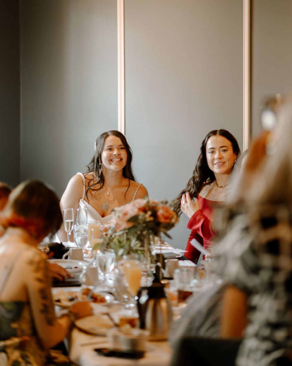Two smiling women in elegant attire seated at a decorated wedding reception table with floral centerpieces.