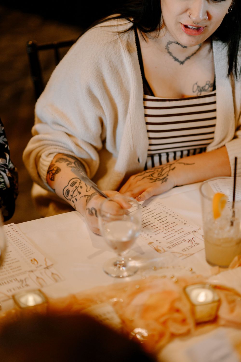 Tattooed woman in striped top and cream cardigan reviewing a menu at a candlelit restaurant table.